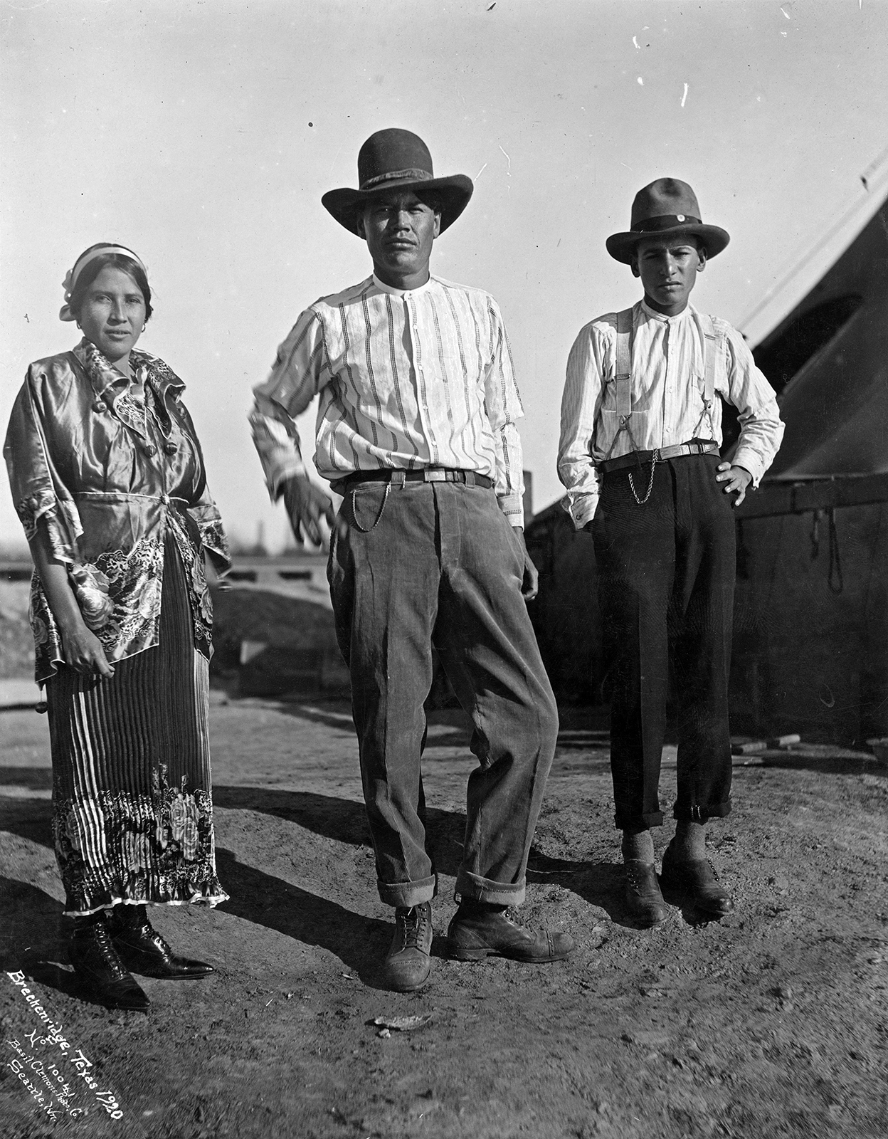 A Mexican woman and two Mexican men, photographed in Breckenridge, Texas, 1920. The three are standing, the woman to the farthest left, and the two men next to her, both men wearing hats. The woman is in a pleated skirt with flower pattern at the bottom, and wears a shiny short-sleeved jacket. 