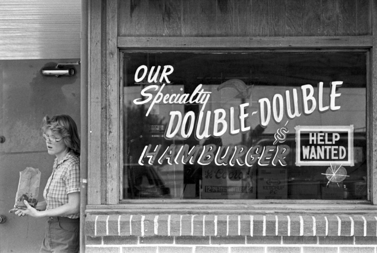 At Al's Drive-In restaurant in Arlington, Missy Freede is seen delivering a bag of burgers alongside an advertisement painted in the store's window for the restaurant's specialty of the Double-Double Hamburger next to a "Help Wanted" sign taped to the window.