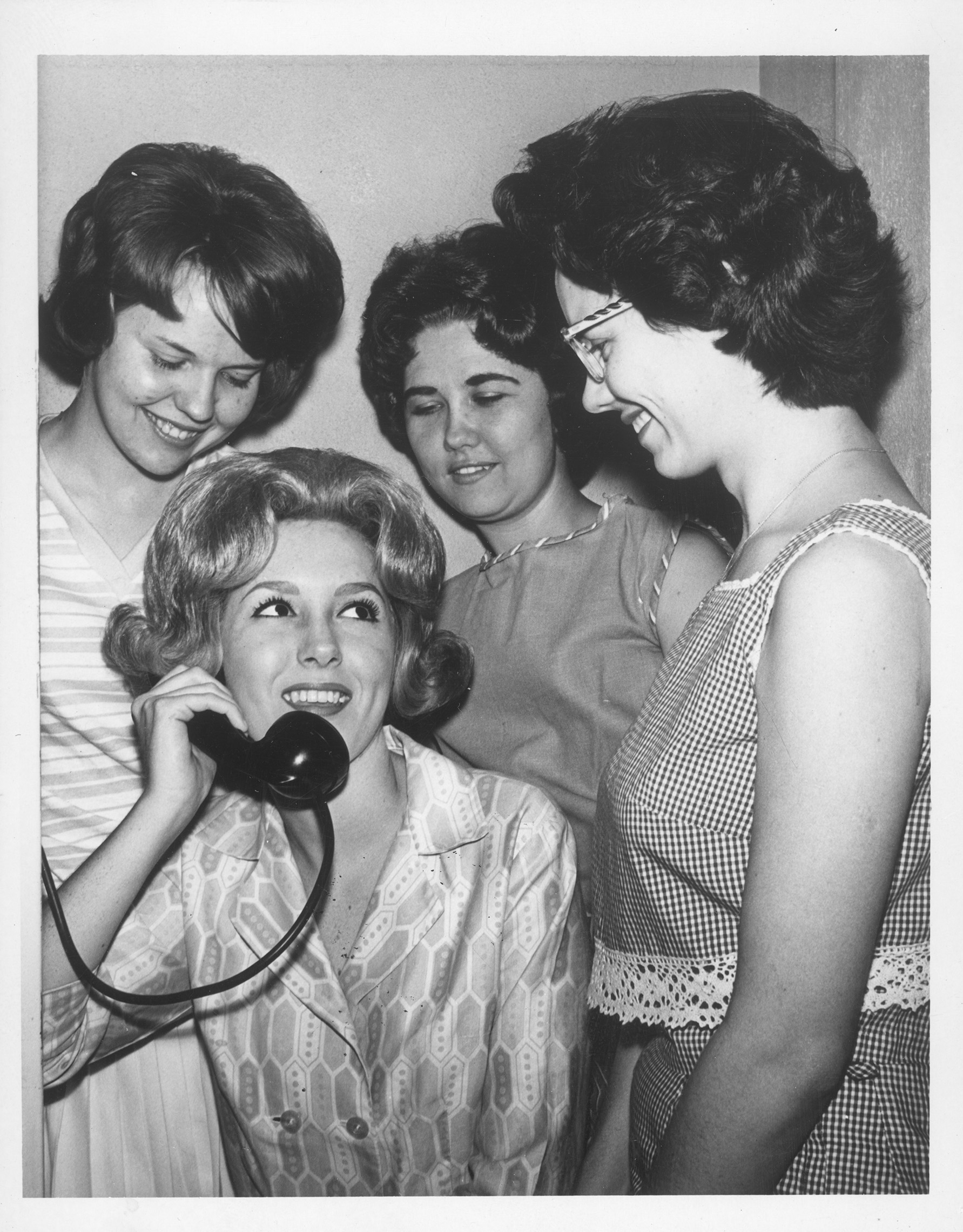 From Arlington State College. Three women stand while one woman speaks into a telephone receiver. Approximately 1950s or 1960s.