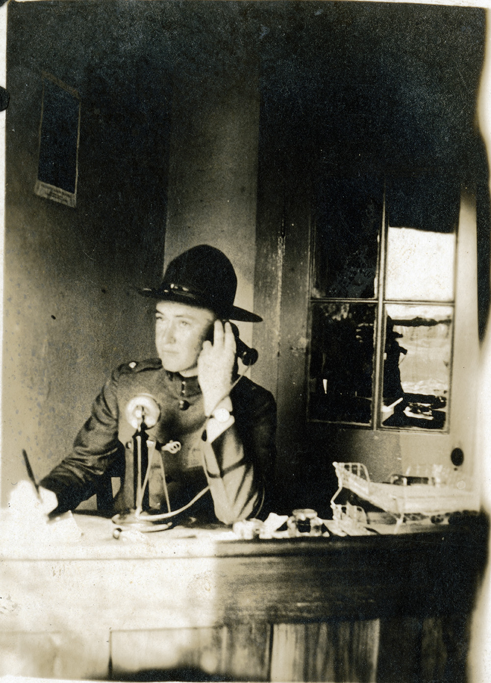  James M. Terrill, wearing a uniform similar to WWI army uniforms, sits at a desk and takes a note while using a telephone from the early 1900's. The telephone has a microphone standing on the desk for the user the speak into, and he is holding up a receiver to his ear to listen to the caller. At Grubbs Vocational College