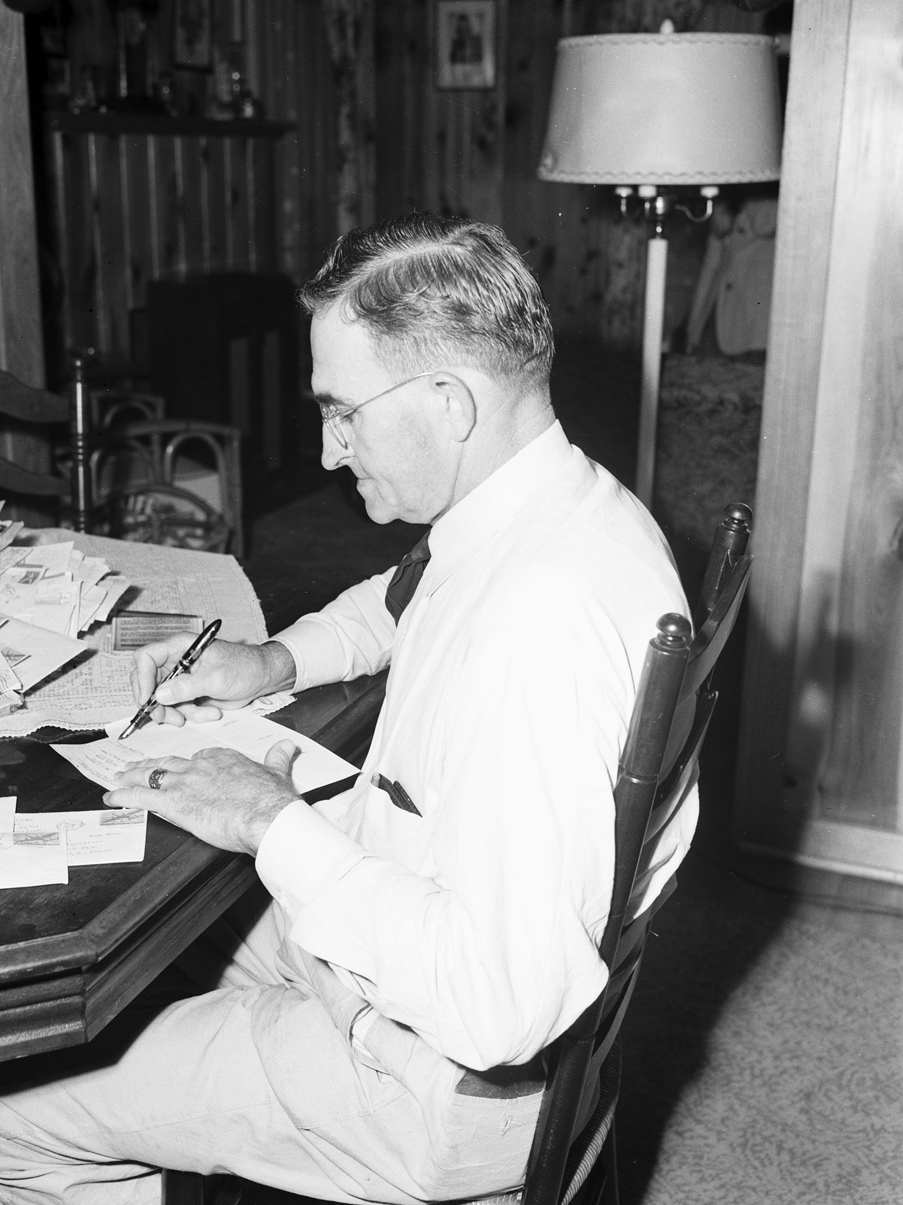 J. J. Boydstun writing letters at his table with a fountain pen. He is wearing a dress shirt, tie, and trousers, and has glasses.