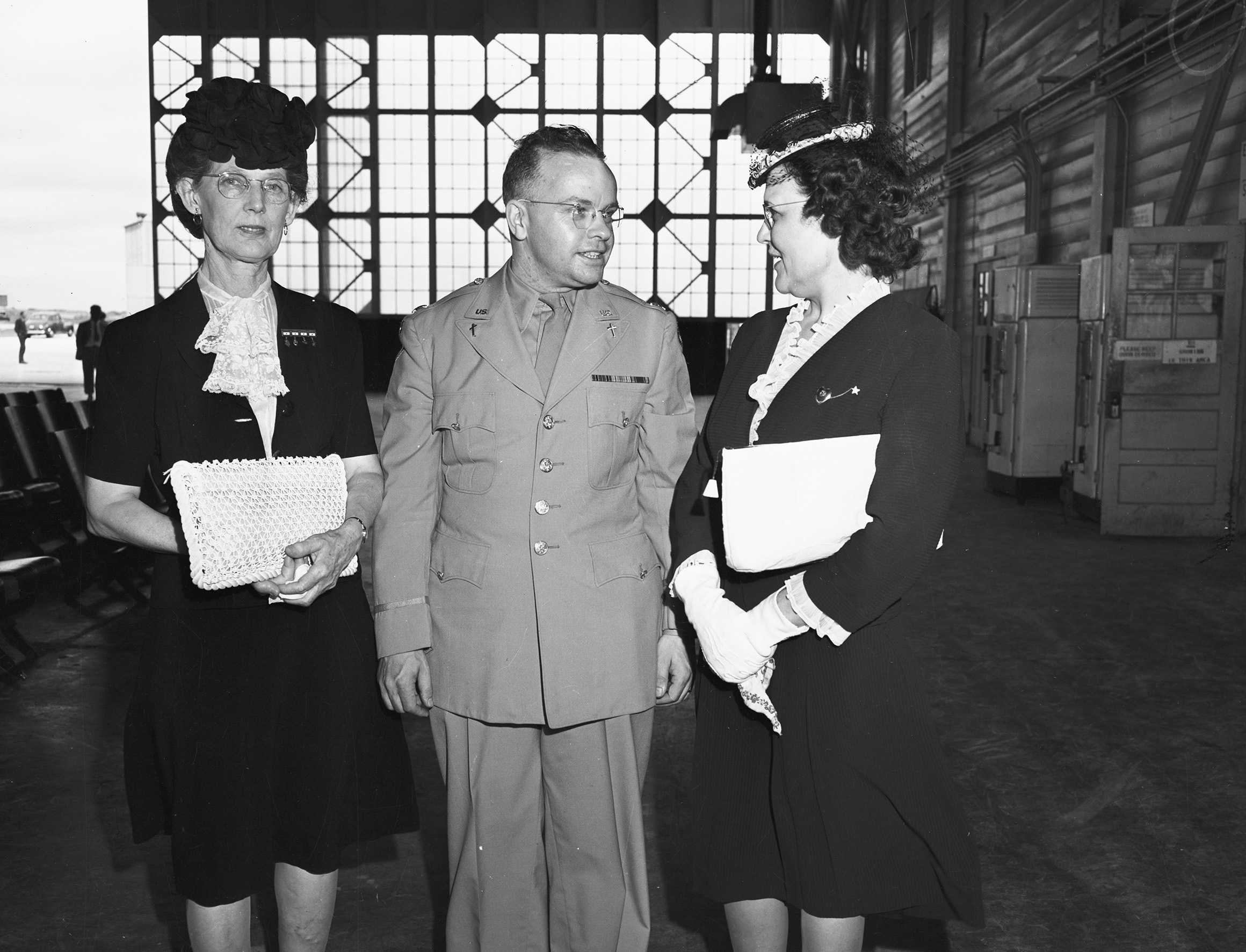Mrs. J. J. Boydstun, left, Chaplain Orville Dennis at center, and Mrs. W. C. O'Briant. The women are wearing dark 1940's styled dressed and Dennis is wearing his military chaplain uniform. They are in what appears to be a plane hangar. 
