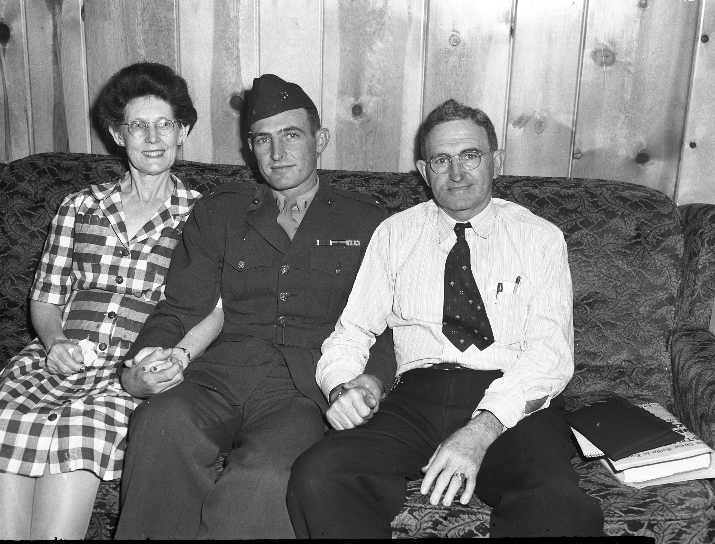 Marine First Lieutenant Robert Boydstun with his parents, Mr. and Mrs. J. J. Boydstun. They are sitting on a couch. His mother is in a checker pattern dress, Robert is at the center in uniform, and his father at right is wearking a dress shirt, tie, and trousers. They are holding hands.