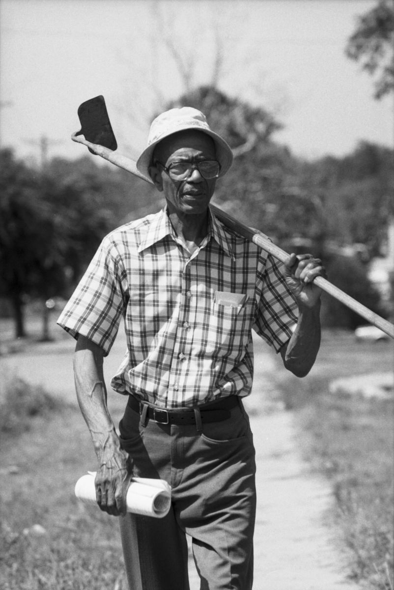 Calvin Littlejohn, pictured with a gardening hoe held over his shoulder and holding a roll of papers.