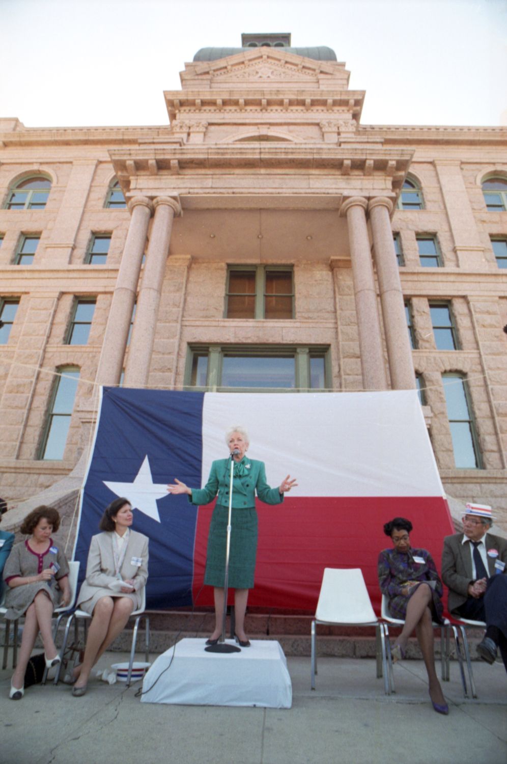 Ann Richards speaking in front of a large Texas flag during a campaign visit to Fort Worth at the Tarrant County Courthouse. The event included a march with supporters from the Tarrant County Courthouse to an absentee voting site at 600 Belknap St.