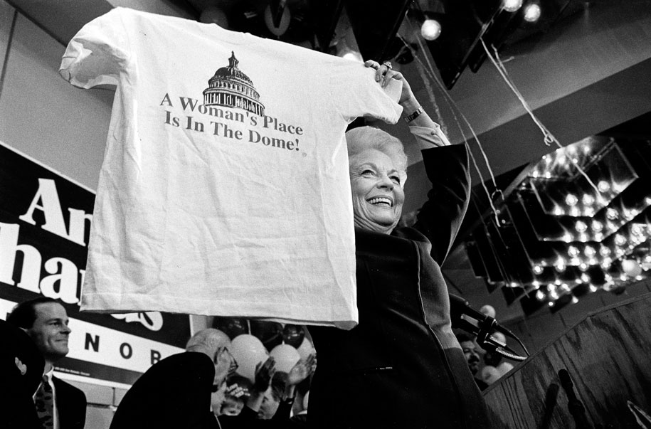 Black and white photograph of Ann Richards holding up a white t-shirt that reads "A Woman's Place is in the Dome!"