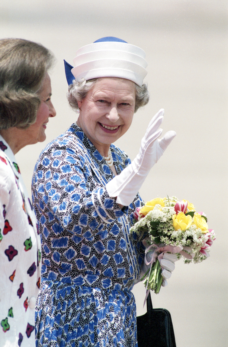 Queen Elizabeth, waving to a crowd (not pictured), with a gloved hand while holding a bouquet of flowers in the other hand.