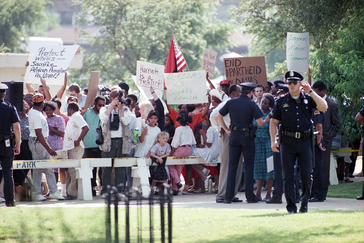 A group of people holding signs and protesting, Dallas Police Officers are seen in the foreground.