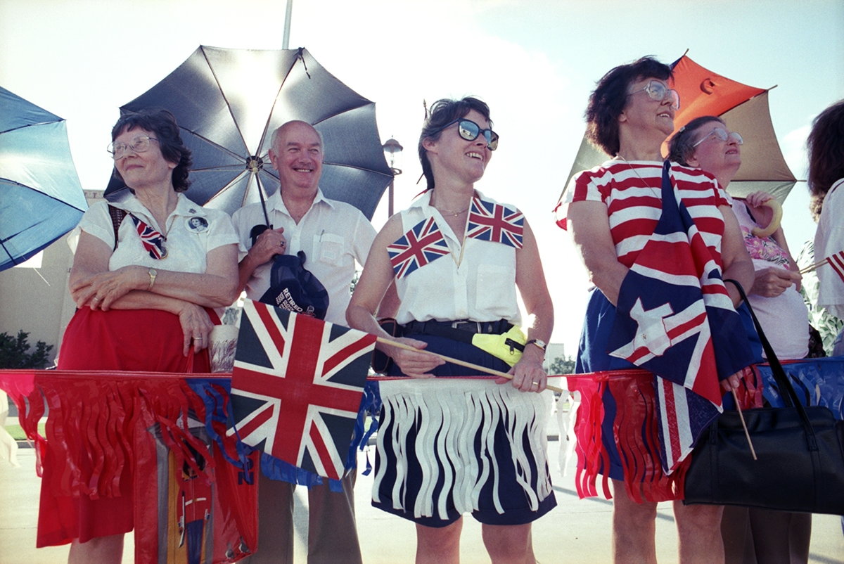 A group of people carrying umbrellas and smiling carrying Union Jack British flags, awaiting the arrival of Queen Elizabeth II at Fair Park in Dallas.