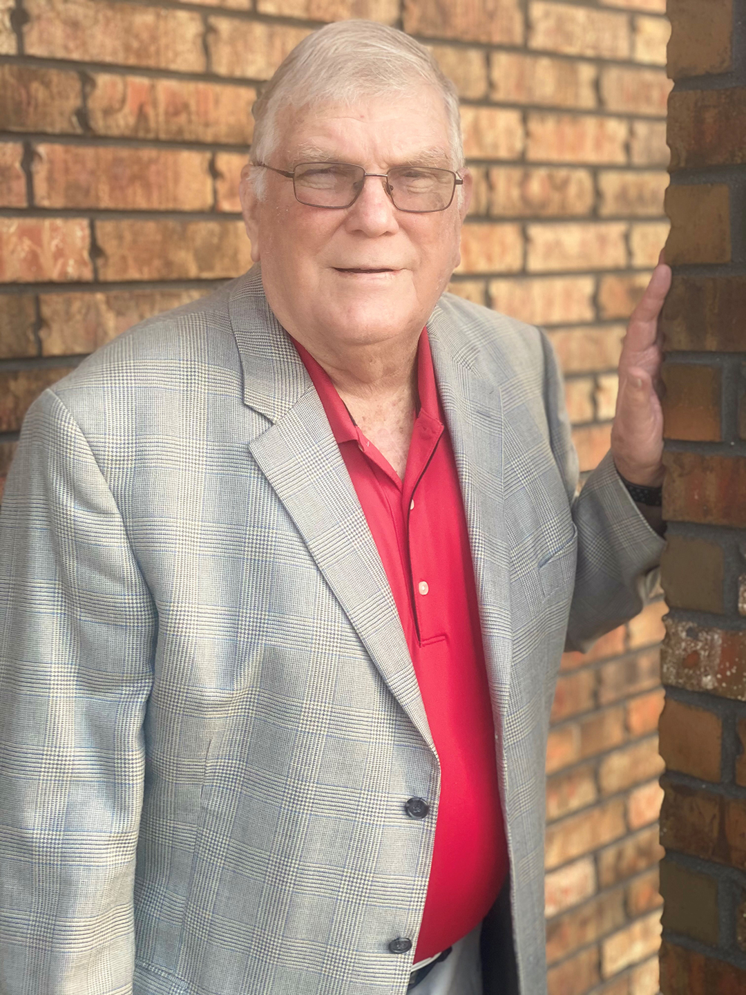 A man in his 80s leans on a brick pillar, smiling at the camera. He is wearing glasses, a red polo, and gray blazer.