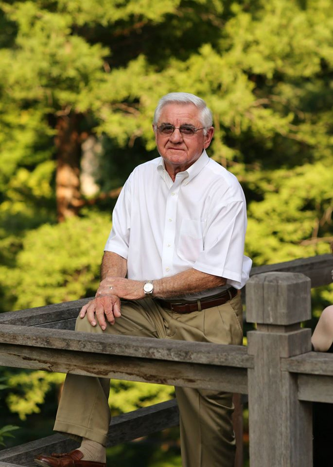 A man in his 80s posts in a natural space, with trees and bright foliage behind him. He is wearing a short sleeve white button down shirt, khakis, and sunglasses.