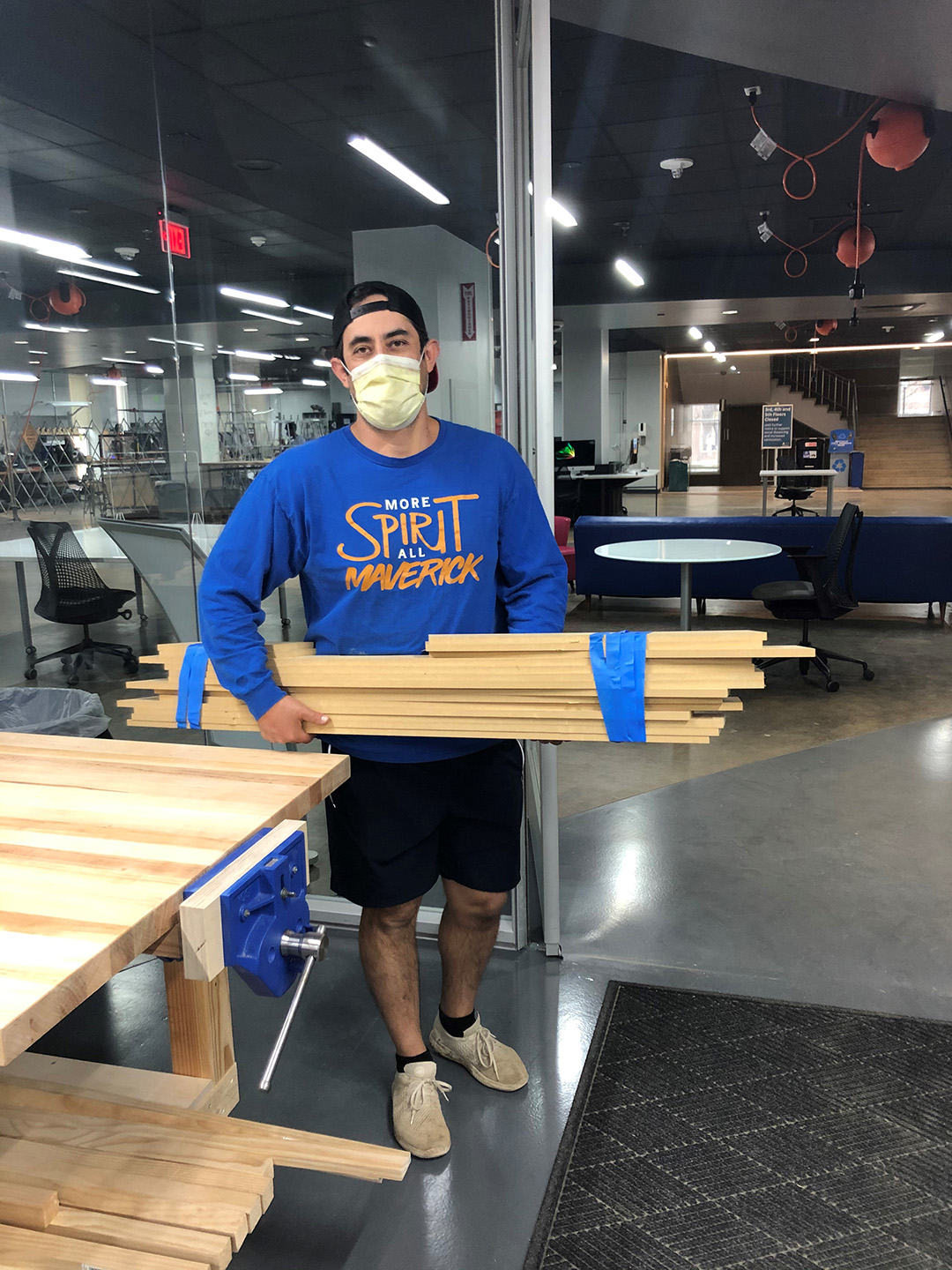a young man stands in the fablab, wearing a face mask and carrying long planks of m d f