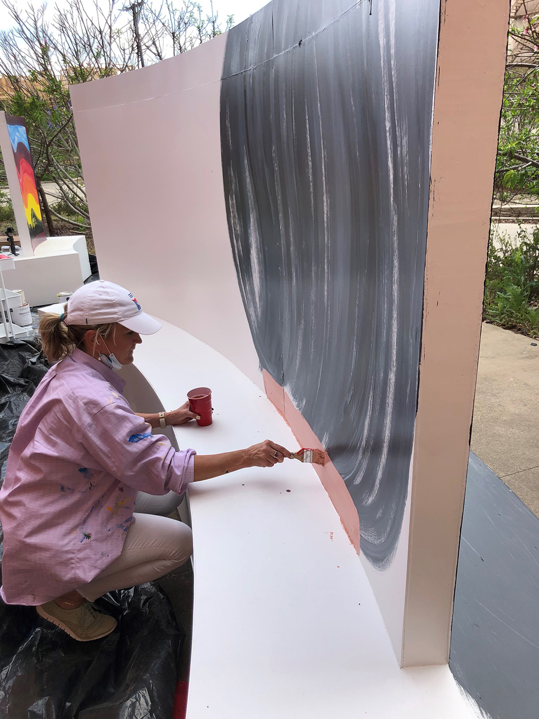 a woman wearing a baseball cap and face mask around her chin painting a large mural on a freestanding wooden structure in an outside area