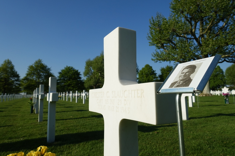 A stone cross adorned for Captain Chester Slaughter, with a photo of him displayed in a metal stand beside it. A large cemetery of hundreds of similar crosses extends around the focal cross.