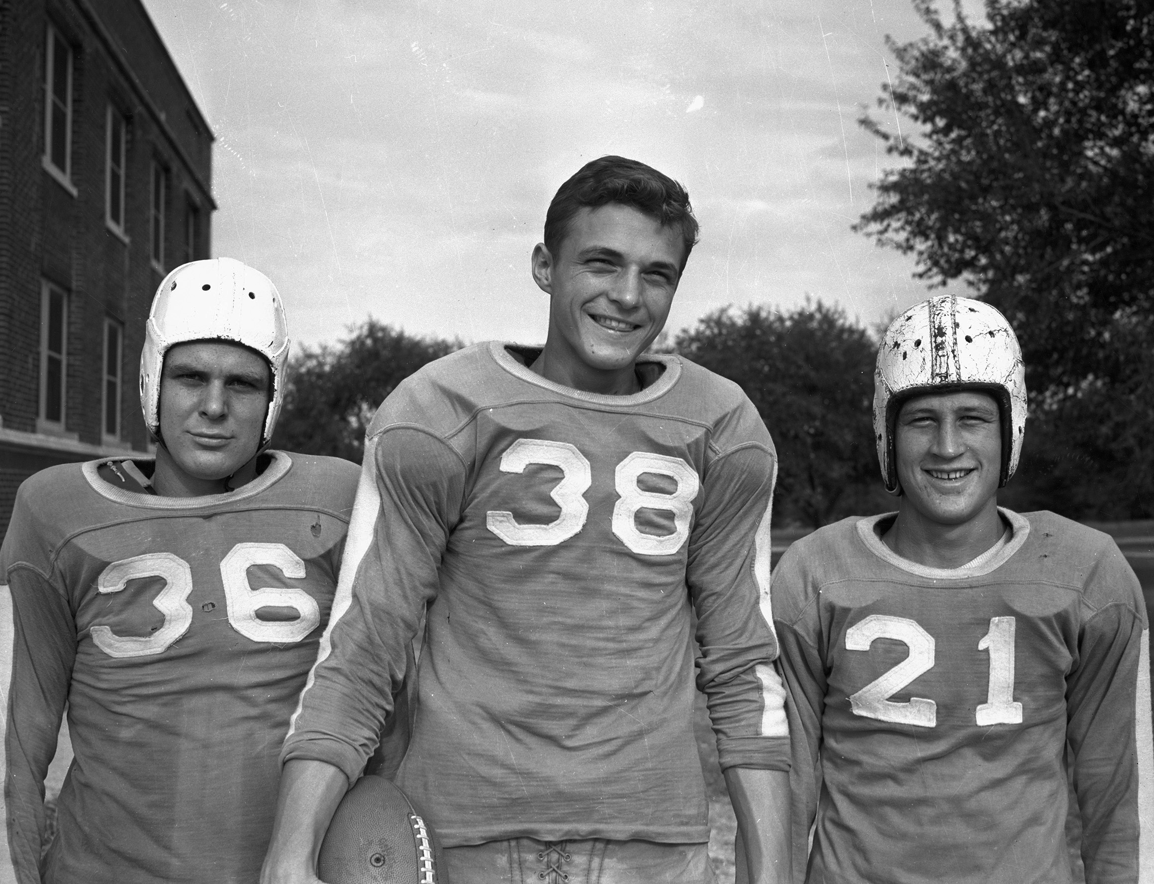 three young men stand side by side in their football uniforms, smiling for the camera; the two on the ends are wearing their helmets, while the middle man is not. behind them are trees and a brick building.
