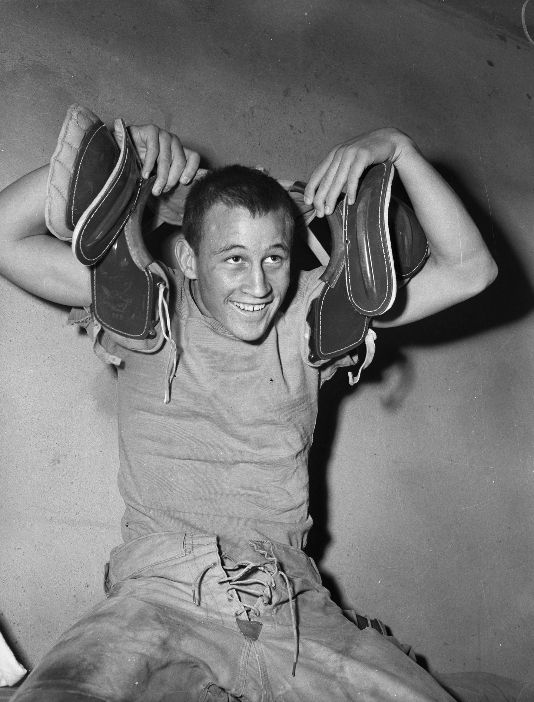A young man is shown dressing in his pads and uniform in the locker room. He is wearing a t-shirt and canvas lace-up pants and holding his shoulder pads in place with a goofy grin on his face. They appear to be held in place with laces.