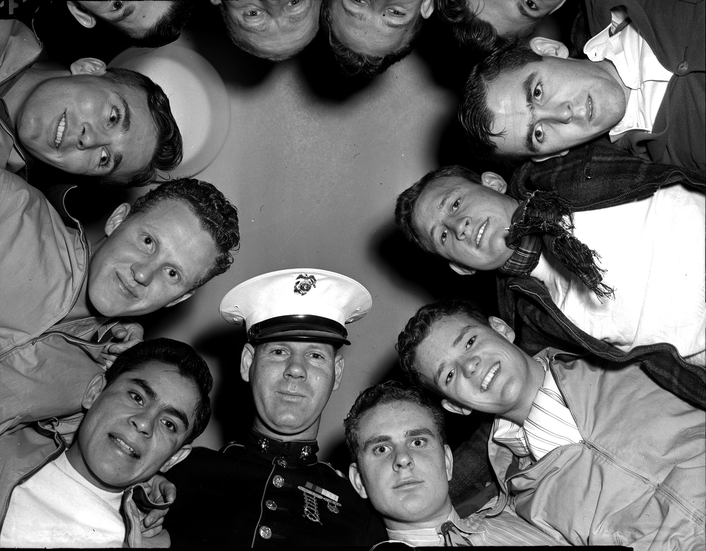 Seven of the ten Masonic Home football players who joined the Marines are shown huddled up with their recruiting officer, Sergeant Fowler. Most of the men are smiling and looking down at the camera. Sergeant Fowler is wearing military uniform.