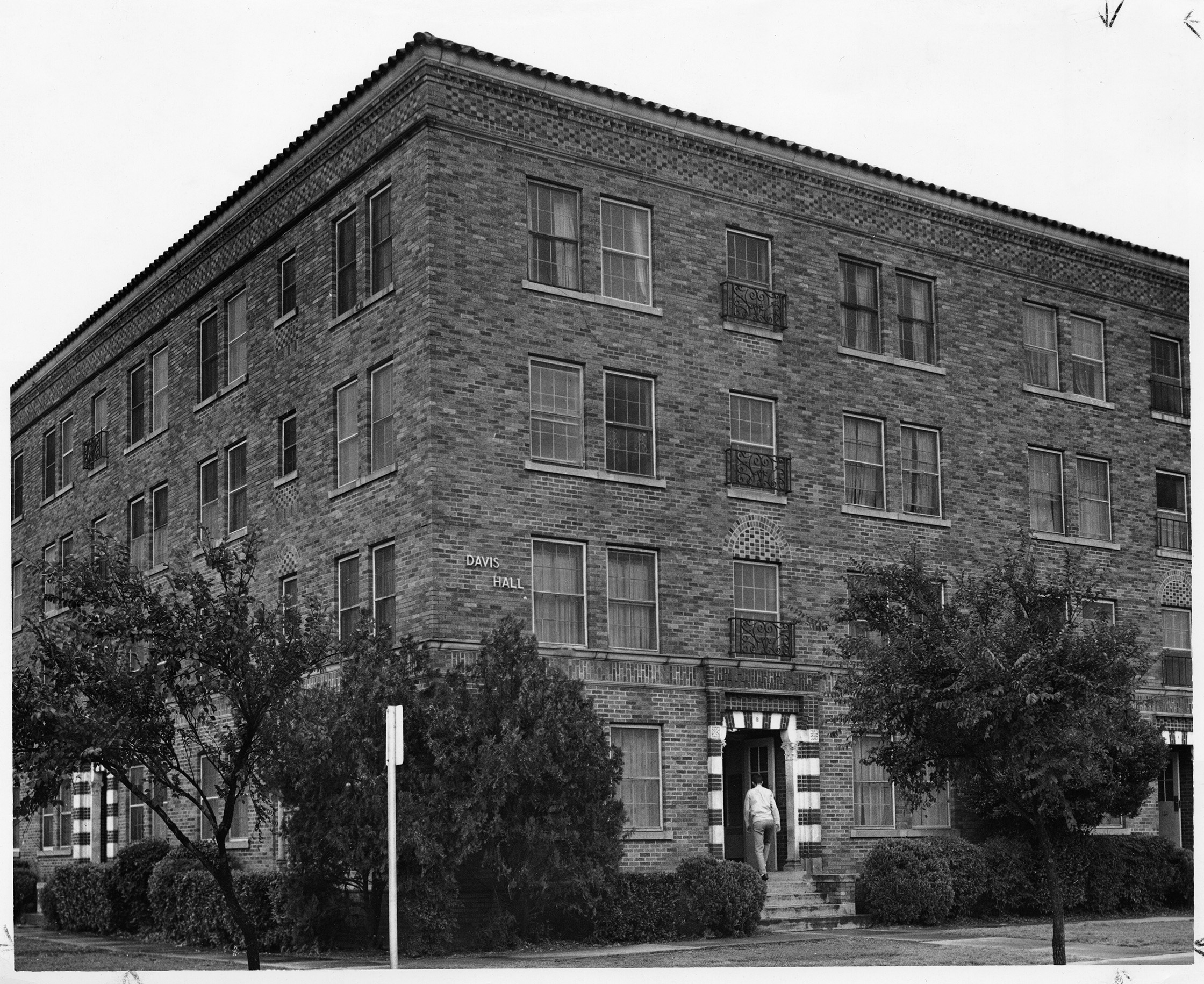 Four-story brick building with trees and shrubs in the foreground. A person is walking into the entrance of the building.