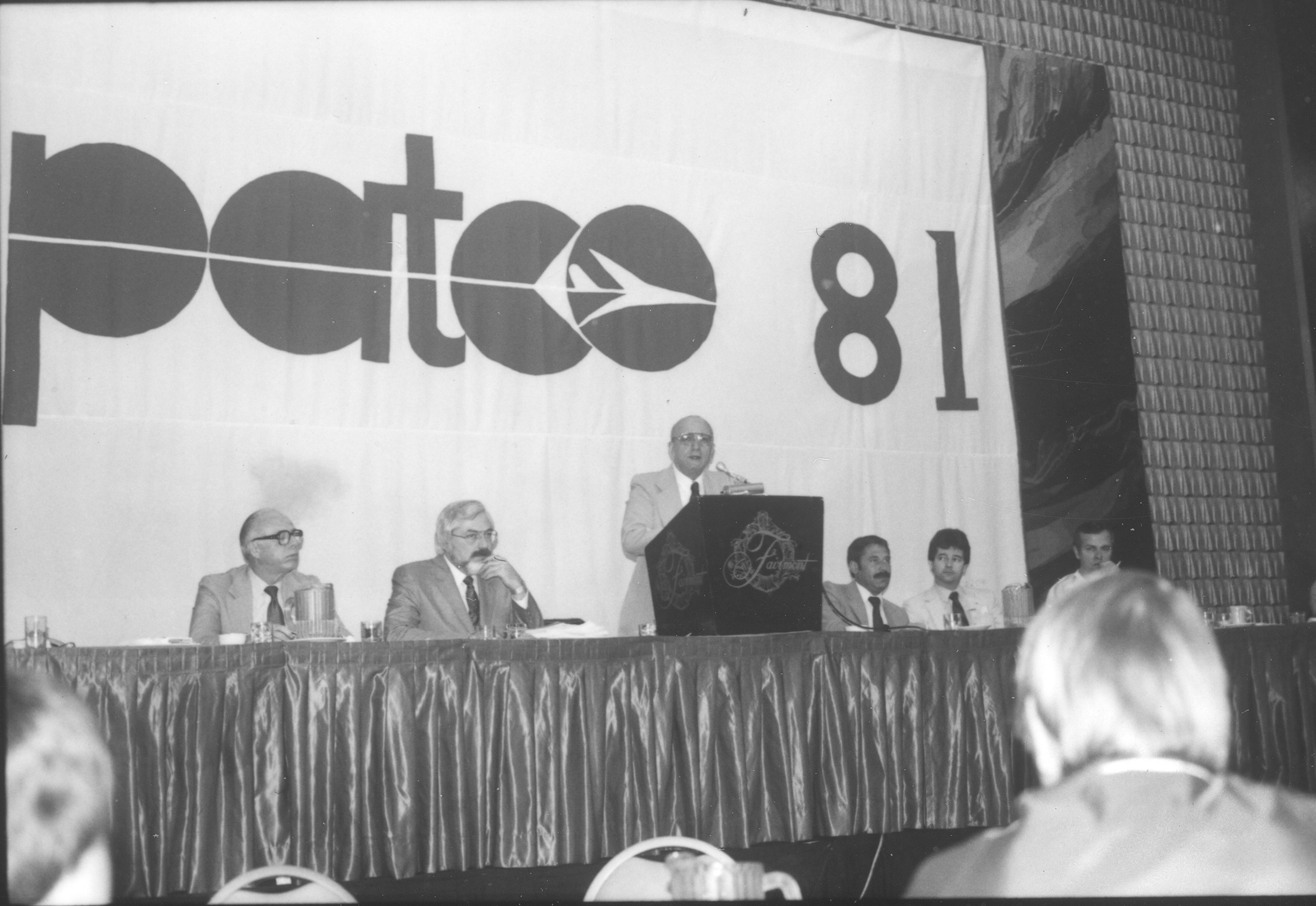 Man speaking at podium with other men alongside him at the presenters' table. "PATCO '81" displayed on wall behind them.