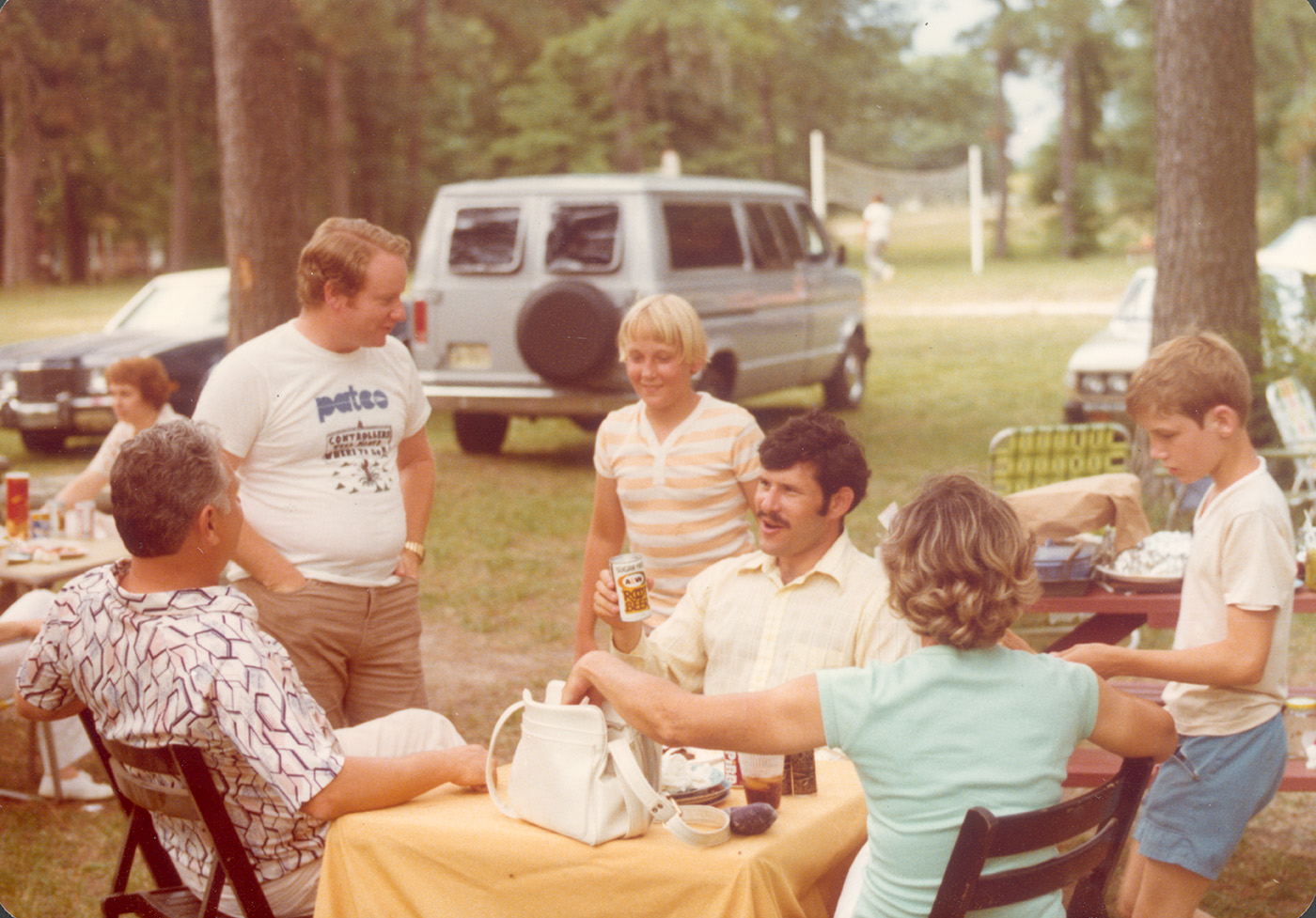 Group of people sitting and standing around a table at a picnic. Cars and trees are in the background.