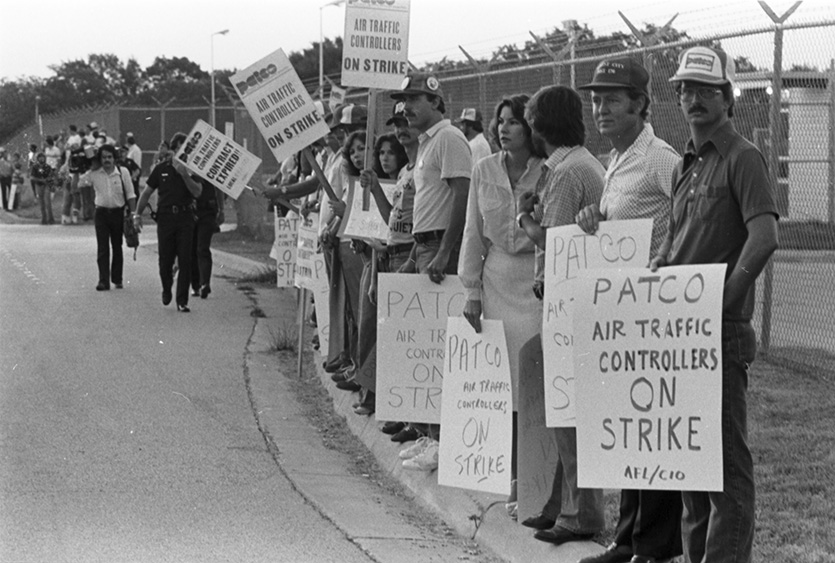 Group of people holding picket signs and standing in a picket line along the curb of a road with a metal fence in the background.