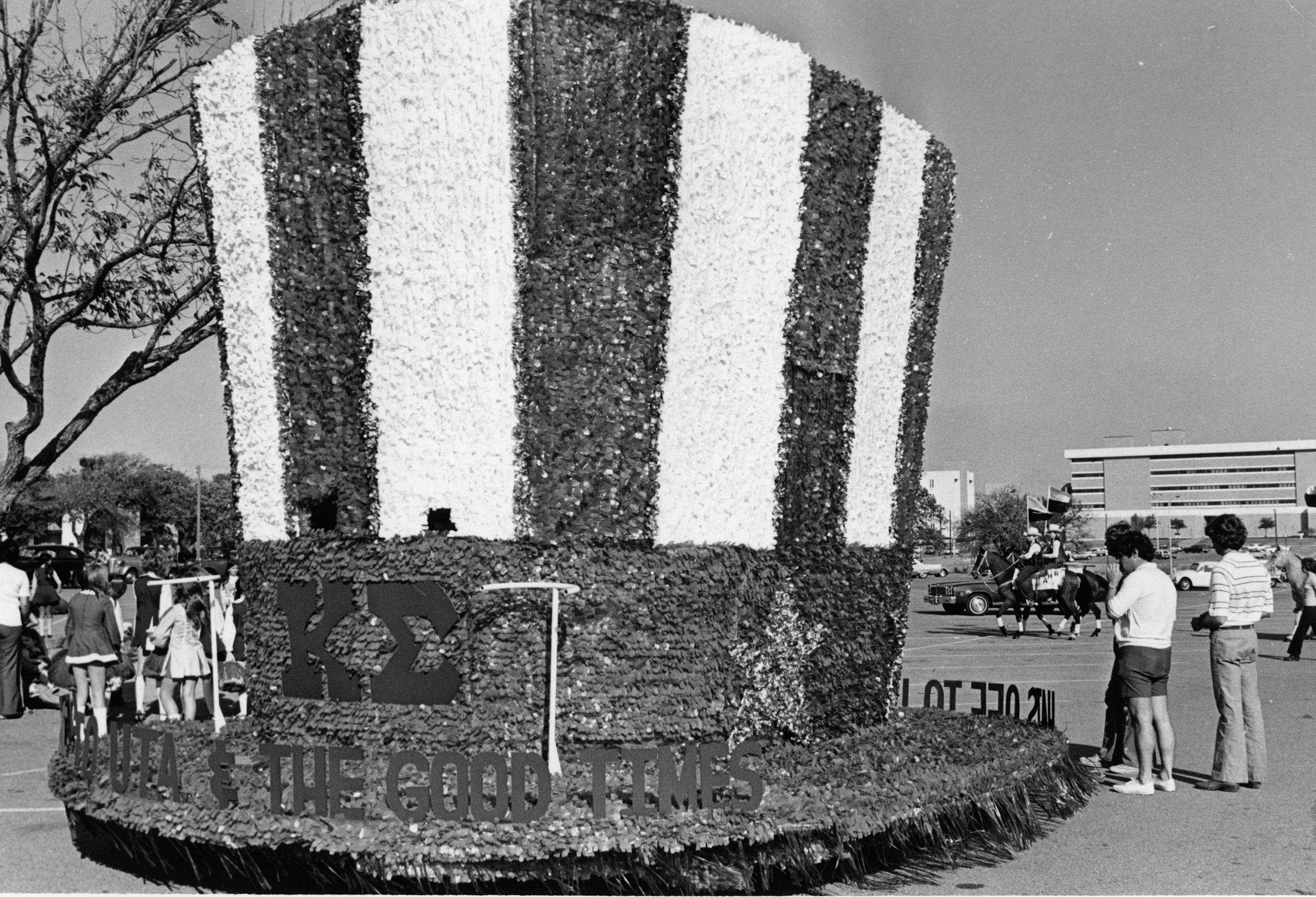 Parade float designed to look like a American flag-style top hat in a parking lot with students, horses, and buildings in the background.