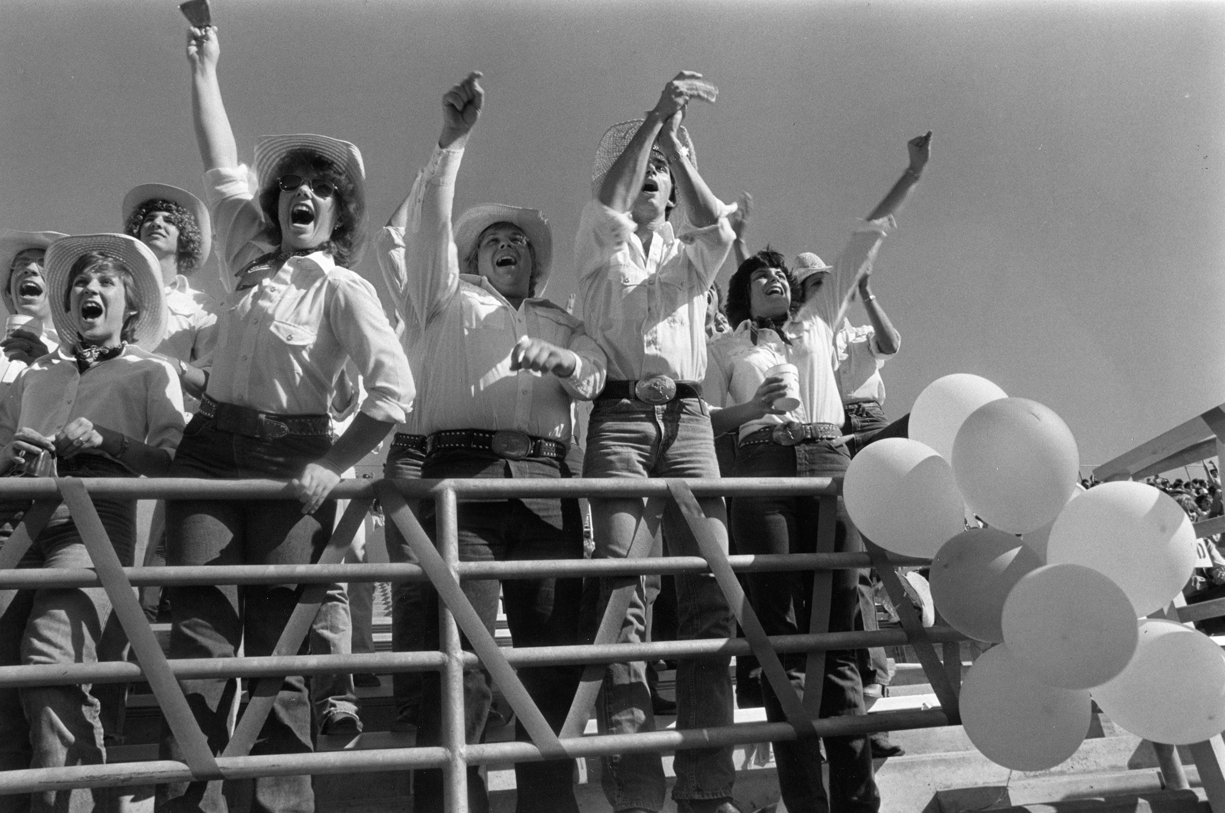 Spirit group cheering and ringing cowbells along the front rail of the stands at a football stadium.