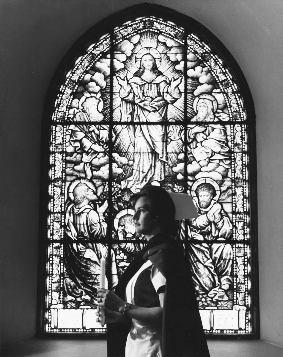 Woman wearing a nurse's uniform and cap holding a candle in front of a stained-glass window