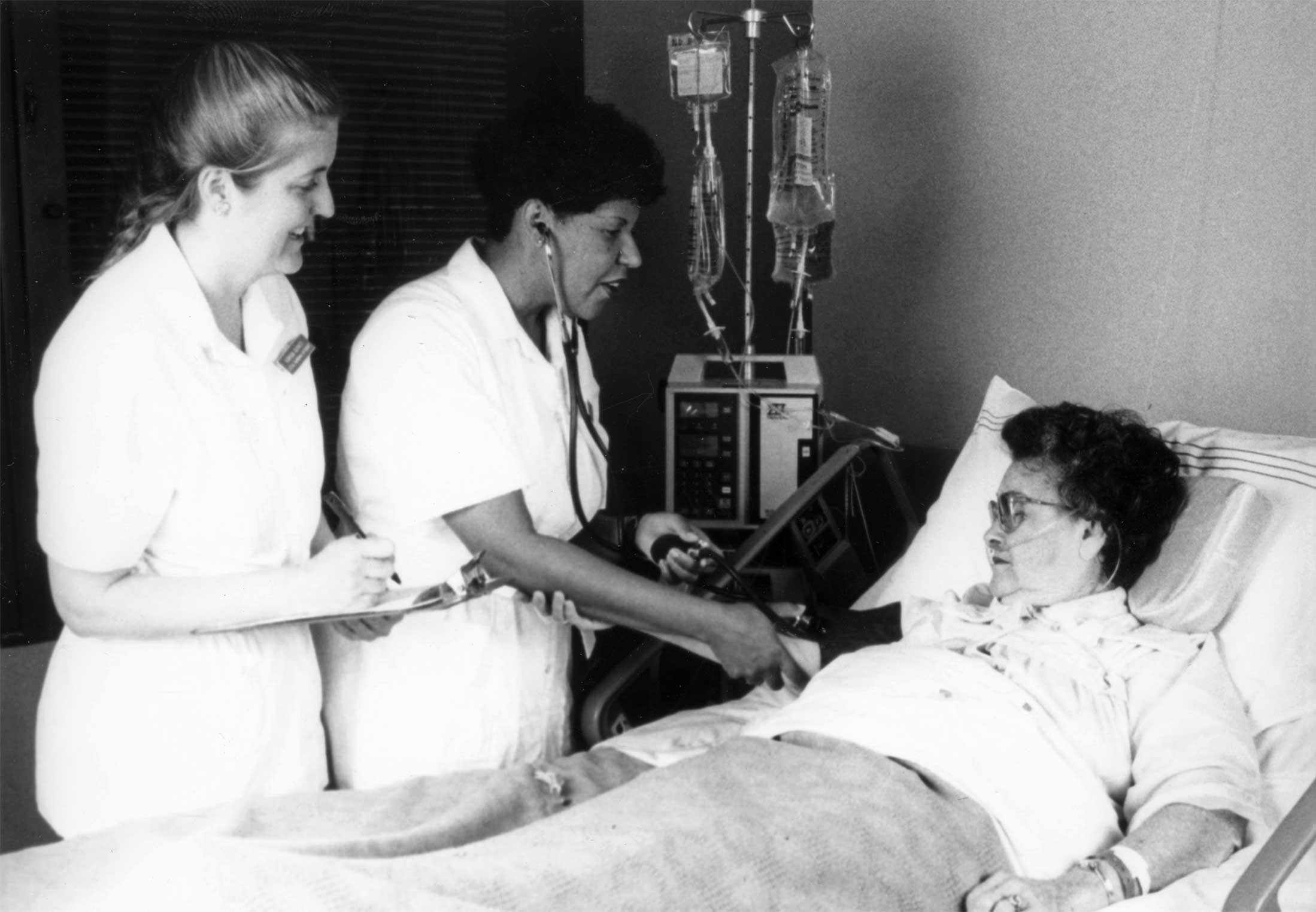 Nurse taking the bloog pressure of a patient lying in a hospital bed while another nurse looks on