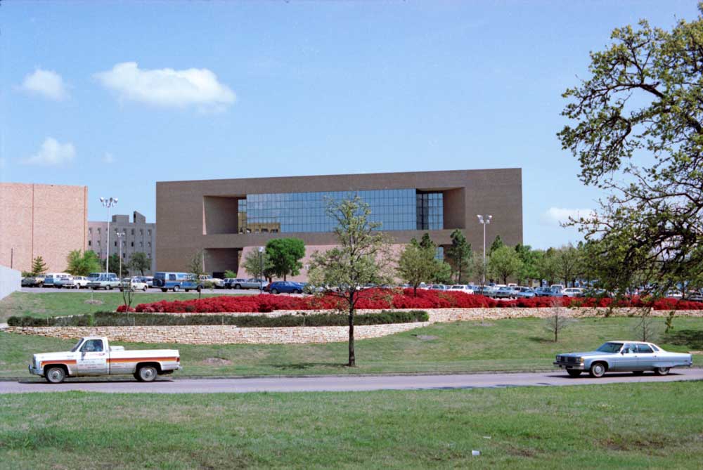 Brick and glass building with landscaping, a parking lot, and a road in front of it