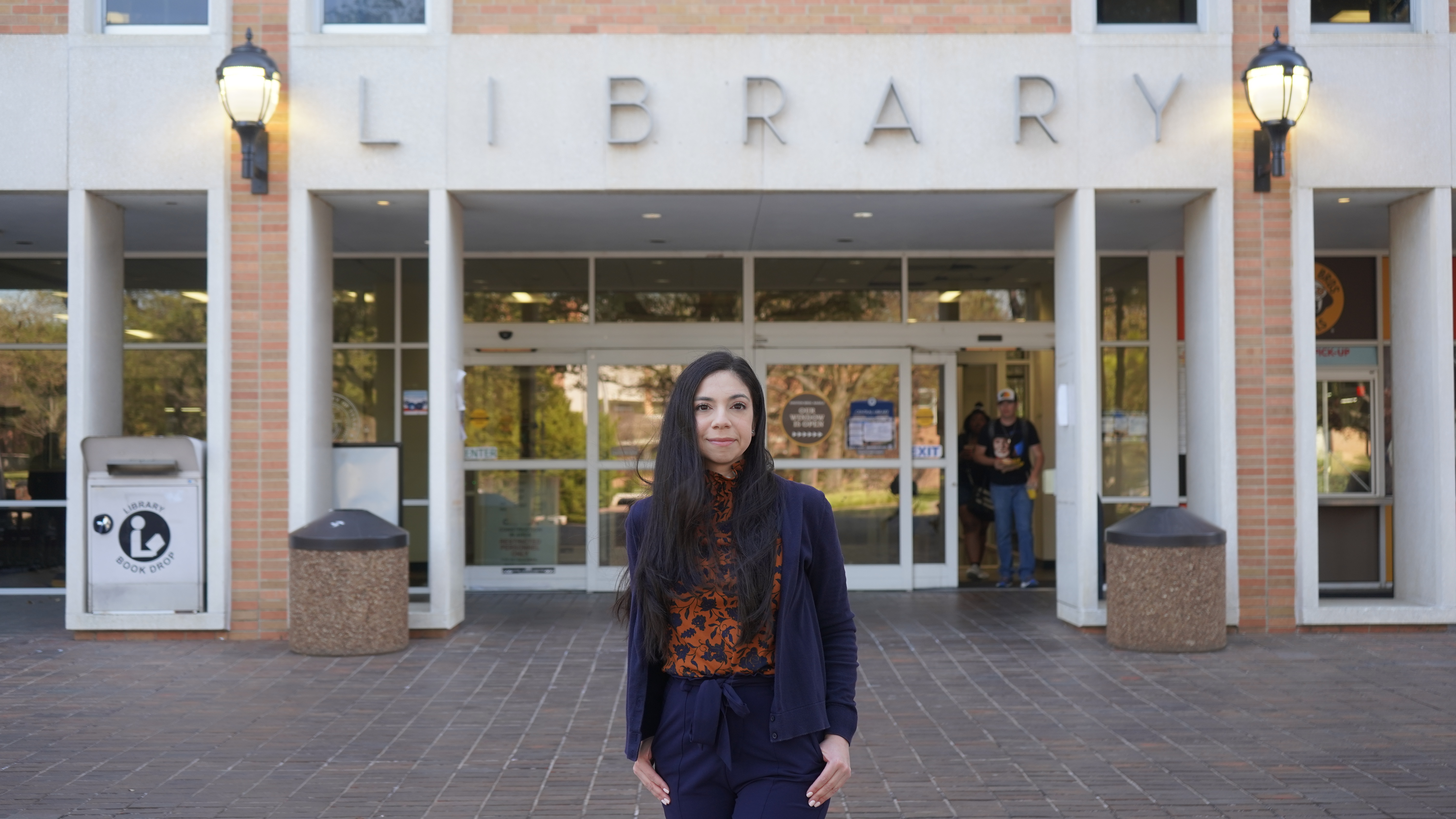 A young woman with long dark hair stands in front of Central Library in a blue tailored suit with an orange patterned top underneath. The word "library" is displayed on the building behind her.