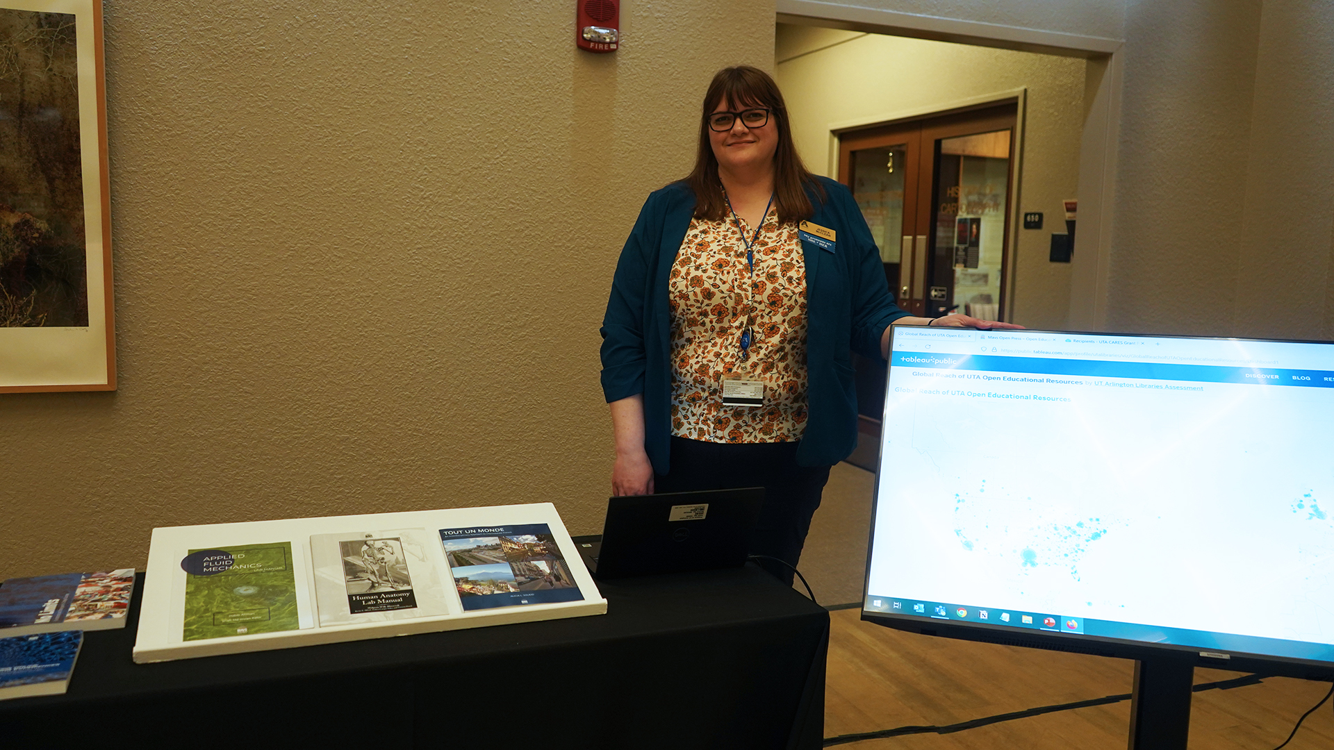 Director of Open Educational Resources Jessica McClean stands beside a digital display of a Tableau and a table display of books in the Atrium of Central Library.