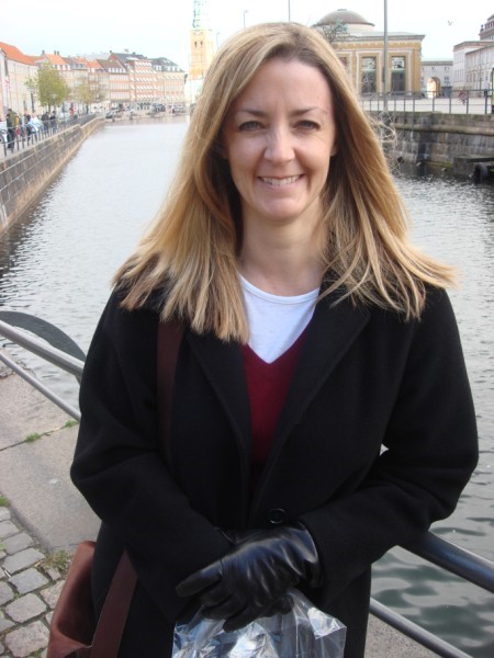 Woman wearing a red and white shirt and a black coat standing in front of a river.