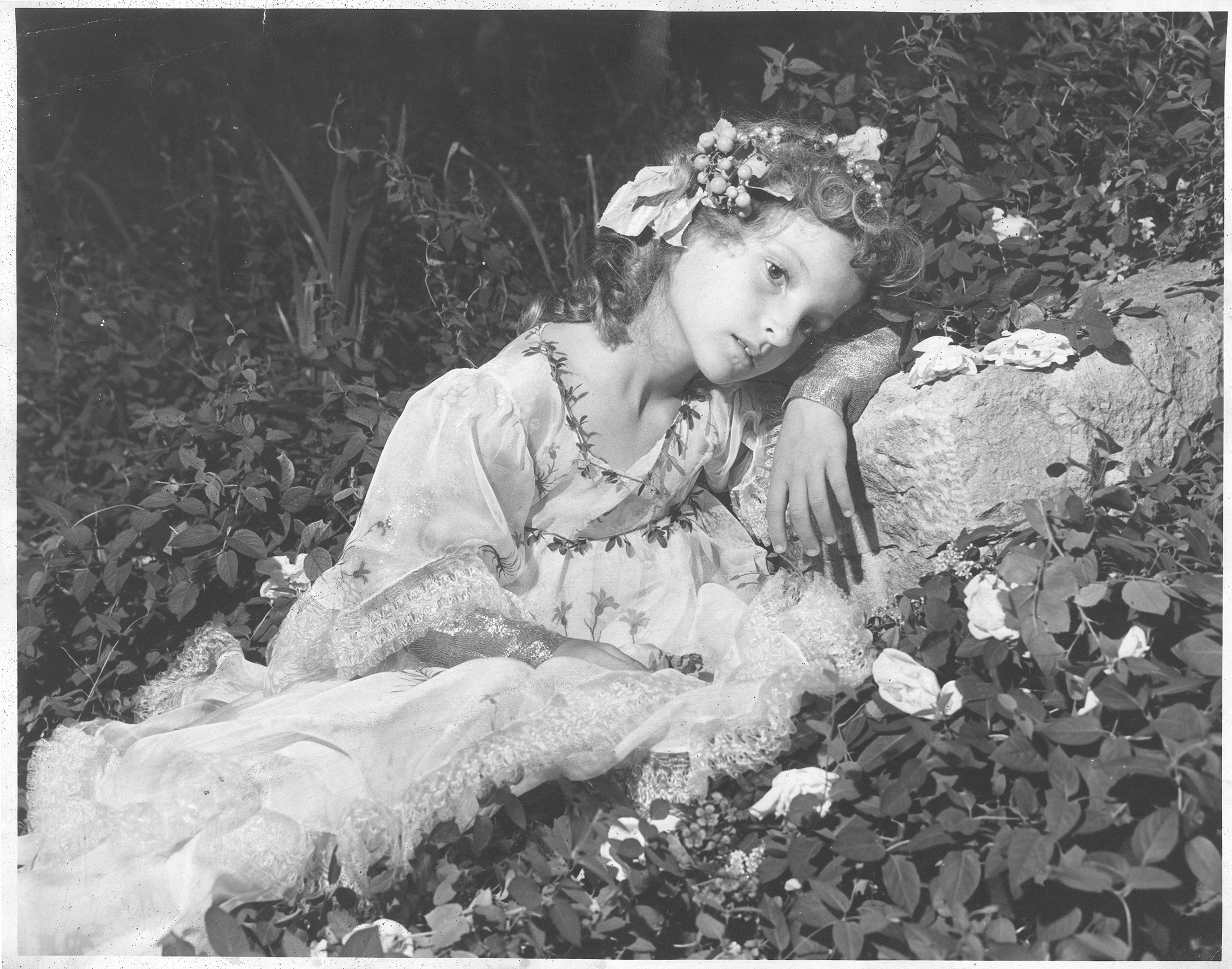 A young girl poses in a bed of foliage wearing a flowing dress and floral headpiece.