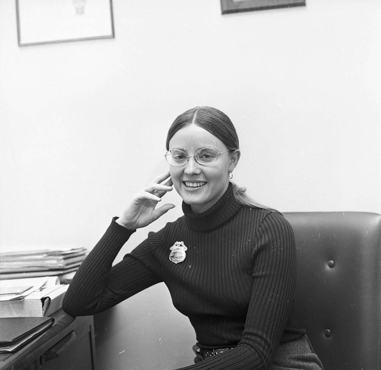 A young woman sits at desk with her elbow on the desk and a police badge on her sweater.