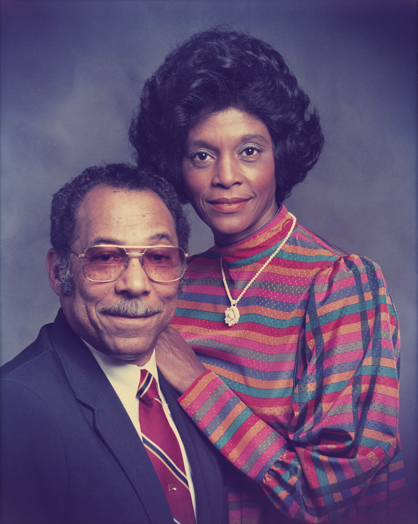 Elzie and Ruby Odom pose against a blue-gray background. Ruby is wearing a brightly patterned top. Elzie has aviator-style glasses.