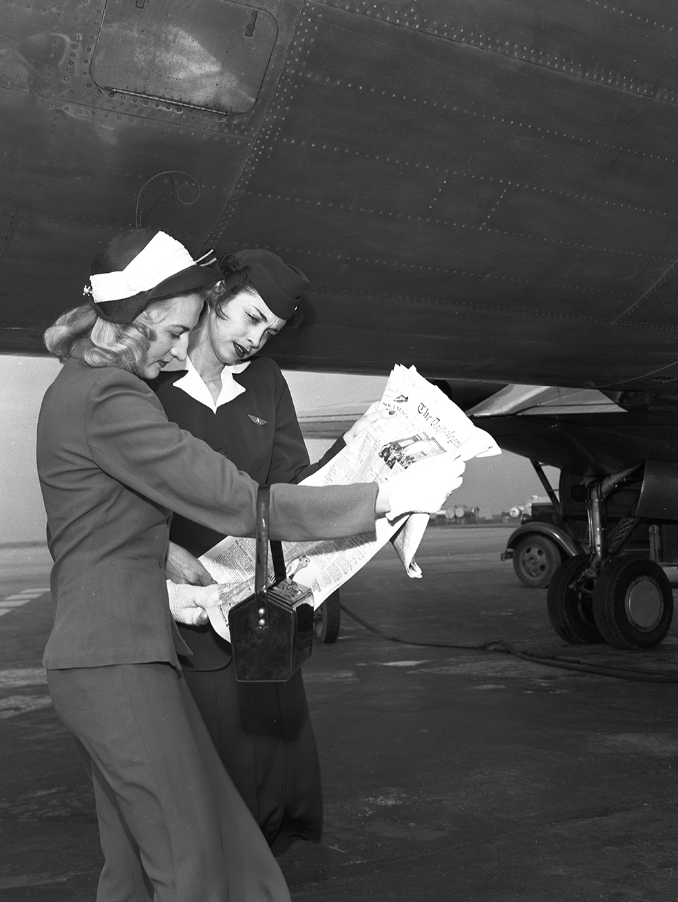 Two young women in tailored jackets and skirts read a newspaper together on a tarmac beside an airplane.