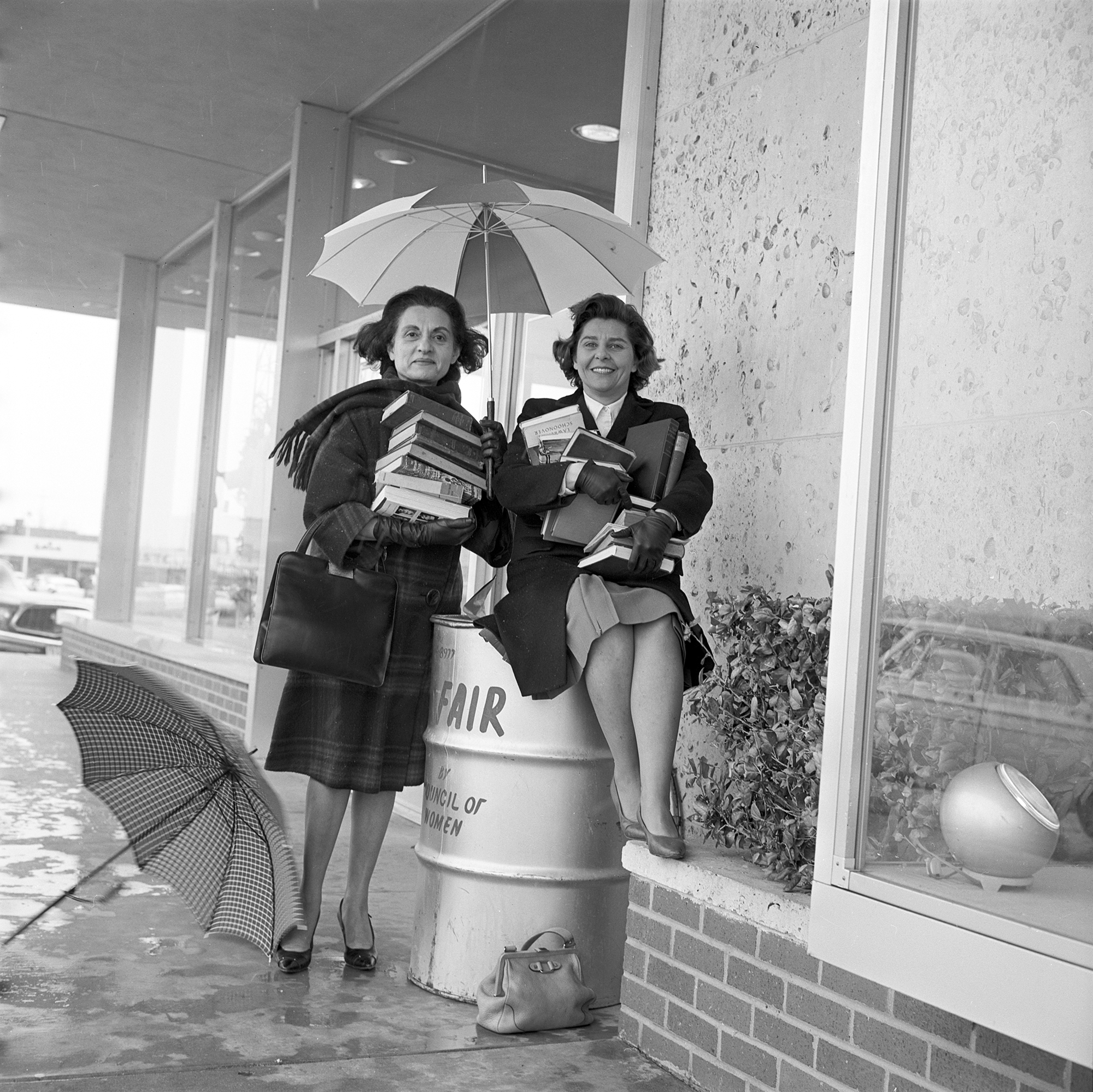 Photo of two women holding large stacks of books while they are positioned under an umbrella and perched on and next to a book barrel.