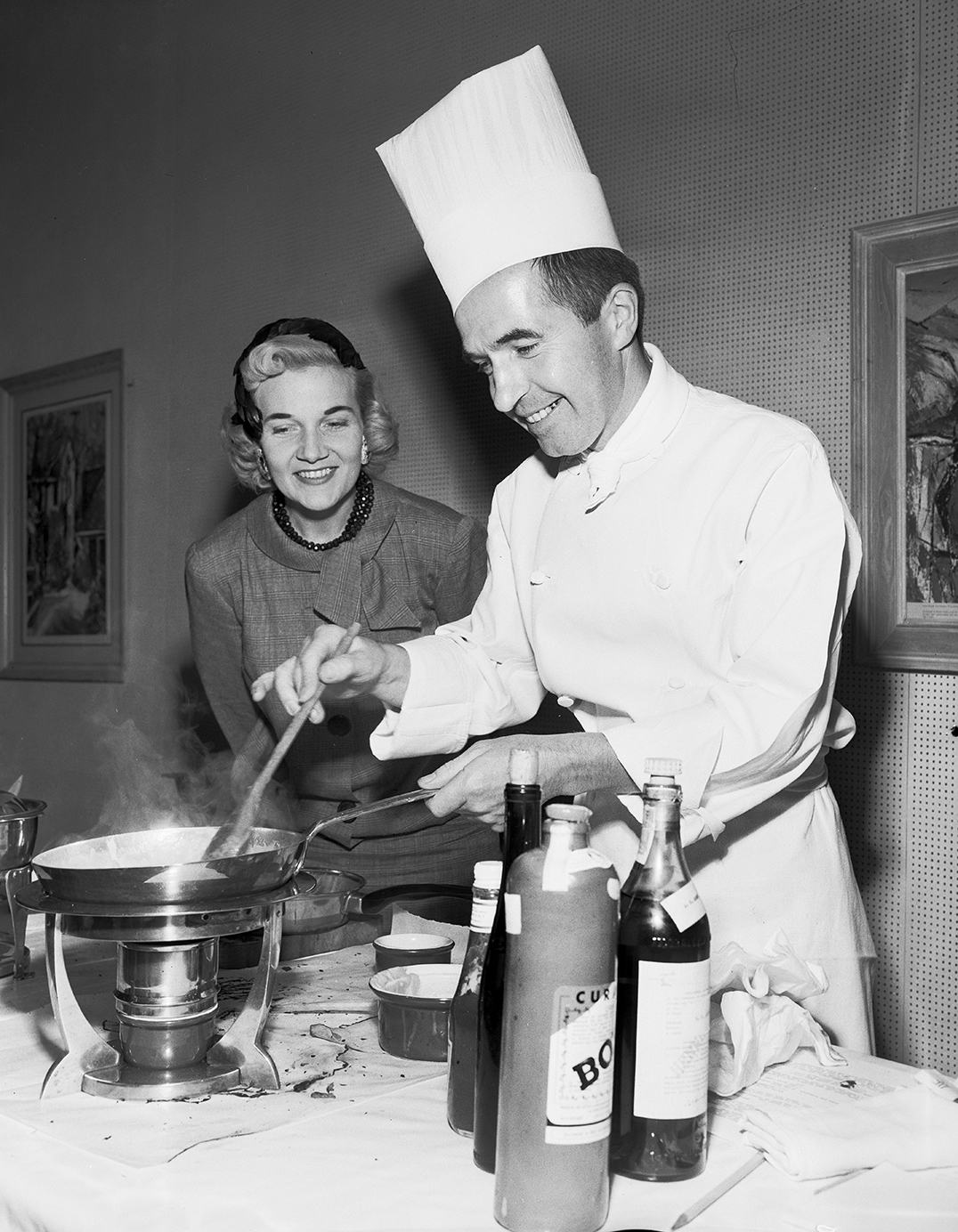 Photo of a chef cooking food in a skillet while a woman looks into the pan from the background.