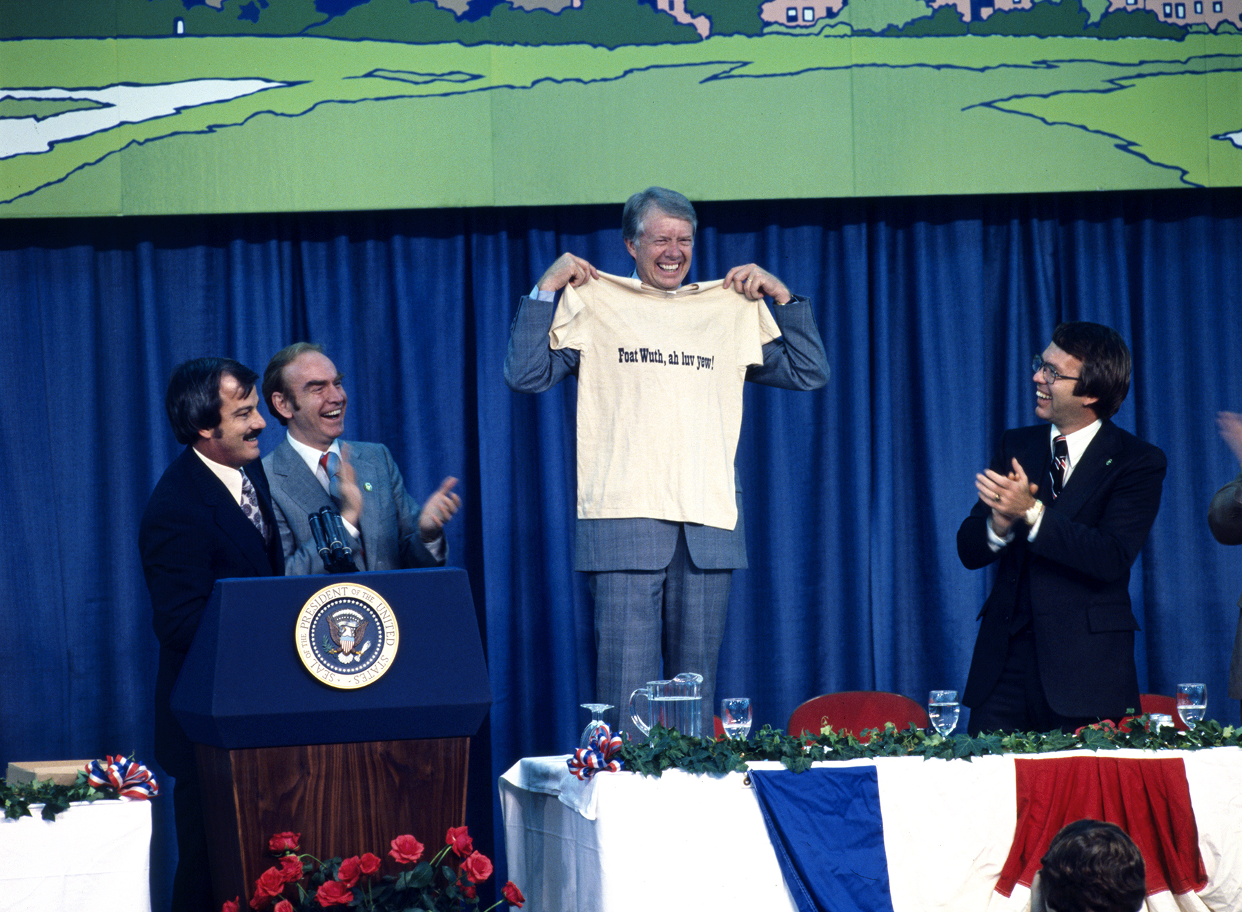 Color photo of President Jimmy Carter holding a shirt reading "Foat Wuth, ah luc yew" while he is smiling and others around him are applauding.