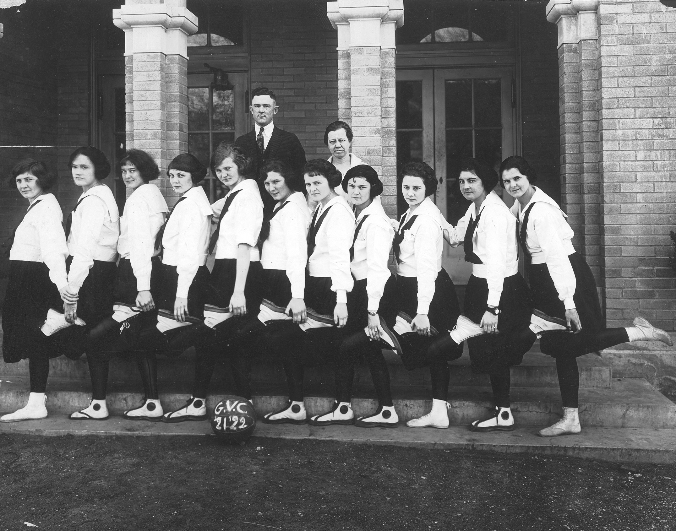 Black and white photo of a group portrait of women on a basketball team, each posed holding the left foot of their teammate. There are a man and woman standing behind the group.