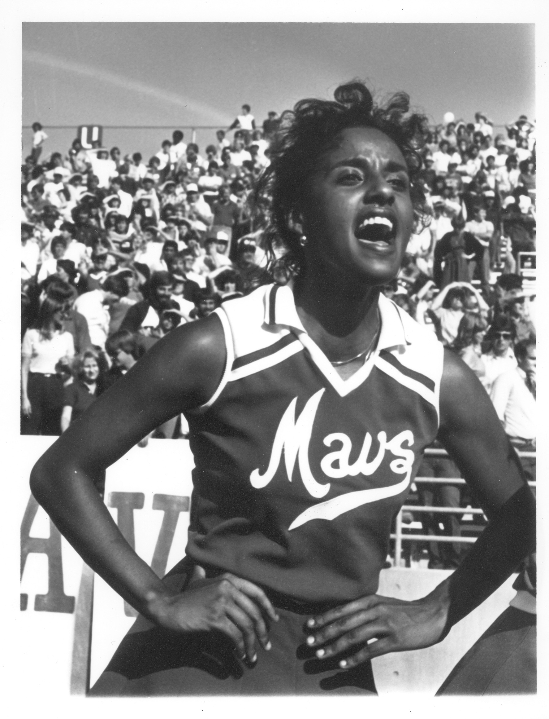 Black and white photo of a cheerleader while cheering at a football game with a large crowd in the stands visible in the background.