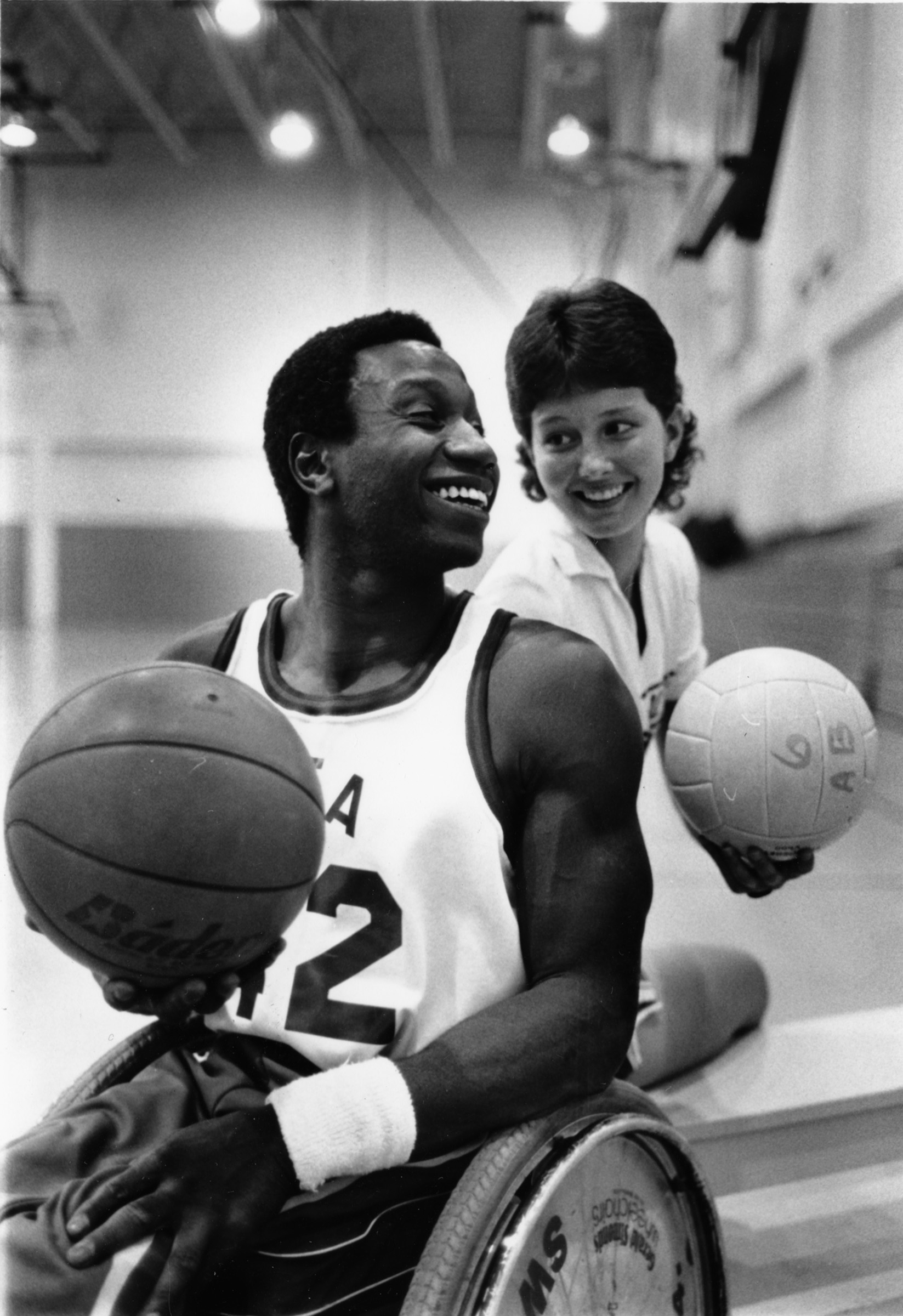 Black and white photo of two people, man on left holding a basketball while seated in a wheelchair, and woman at right holding a volleyball. They are looking at each other.