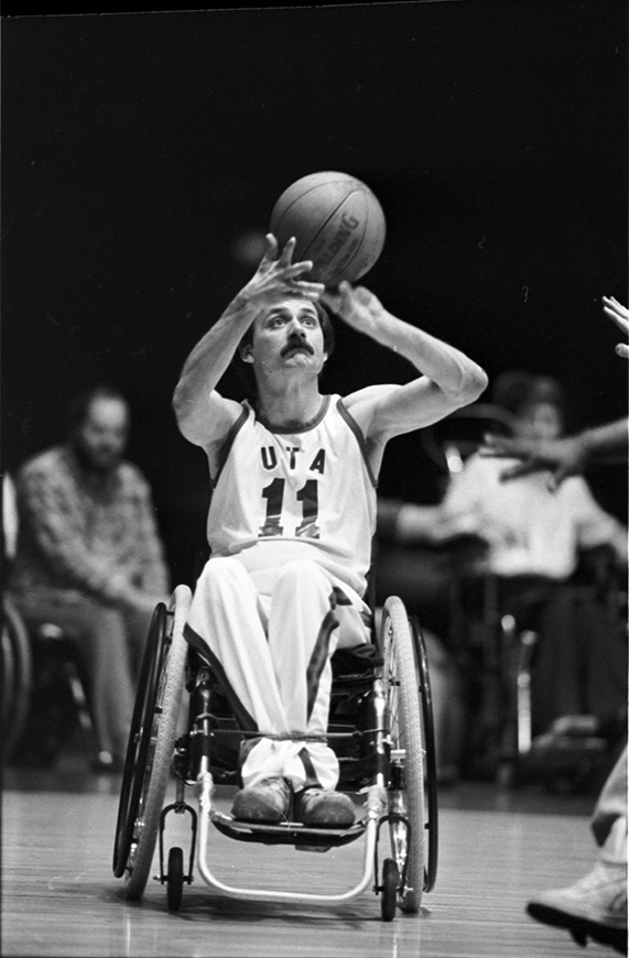 Black and white photo of a wheelchair user taking a shot with a basketball during a wheelchair basketball game.