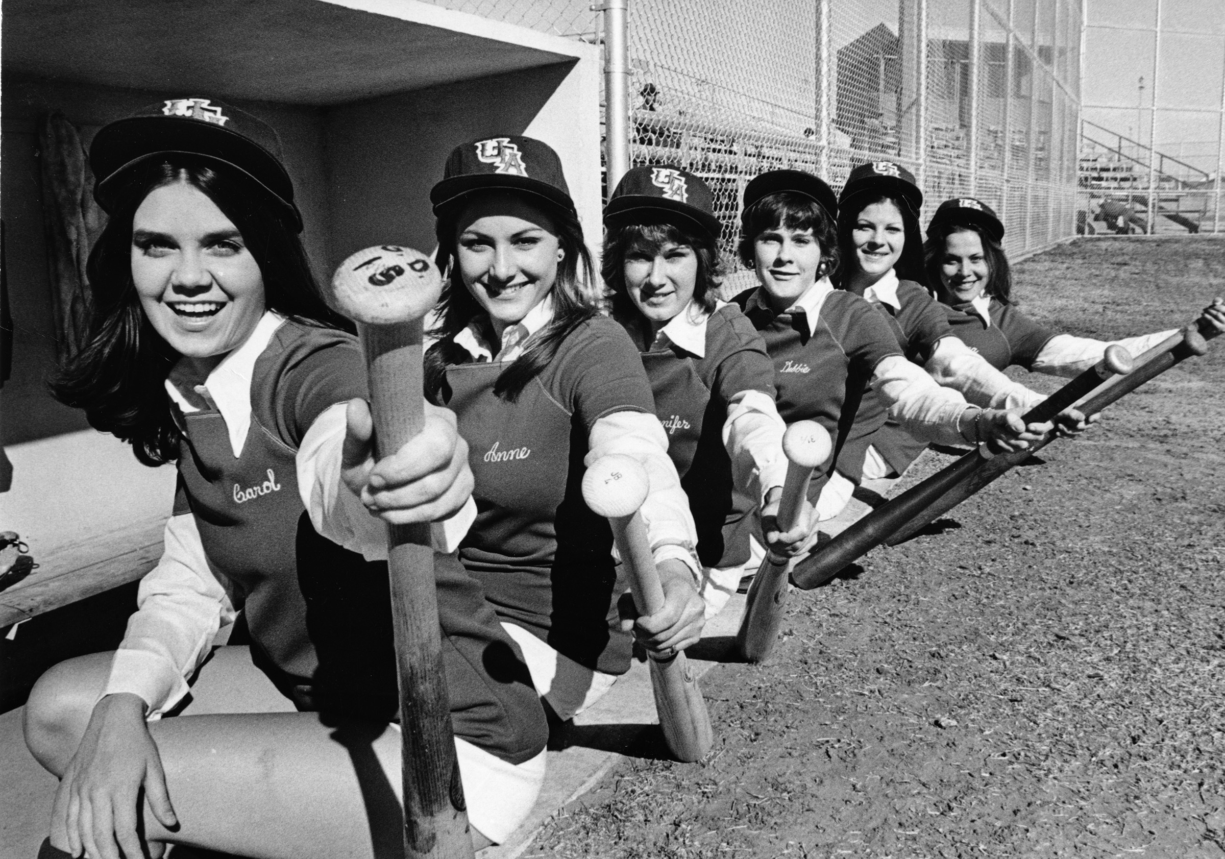 Black and white photo of a group of softball players posed sitting in a dugout holding bats and looking towards the camera.