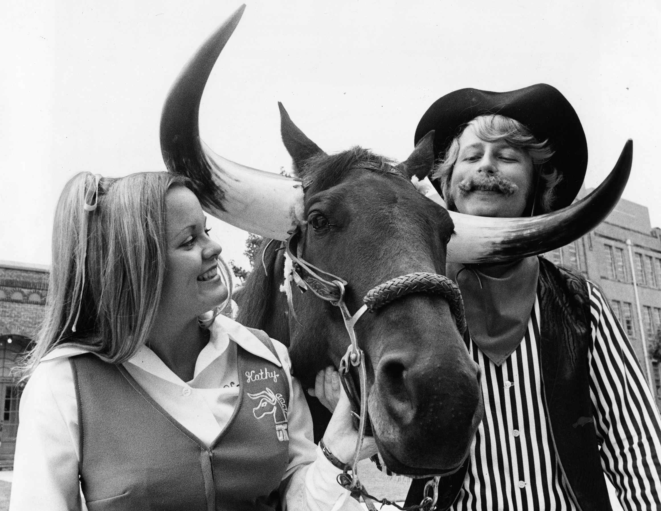 Black and white photo of two people and a horse with faux horns attached to its head.
