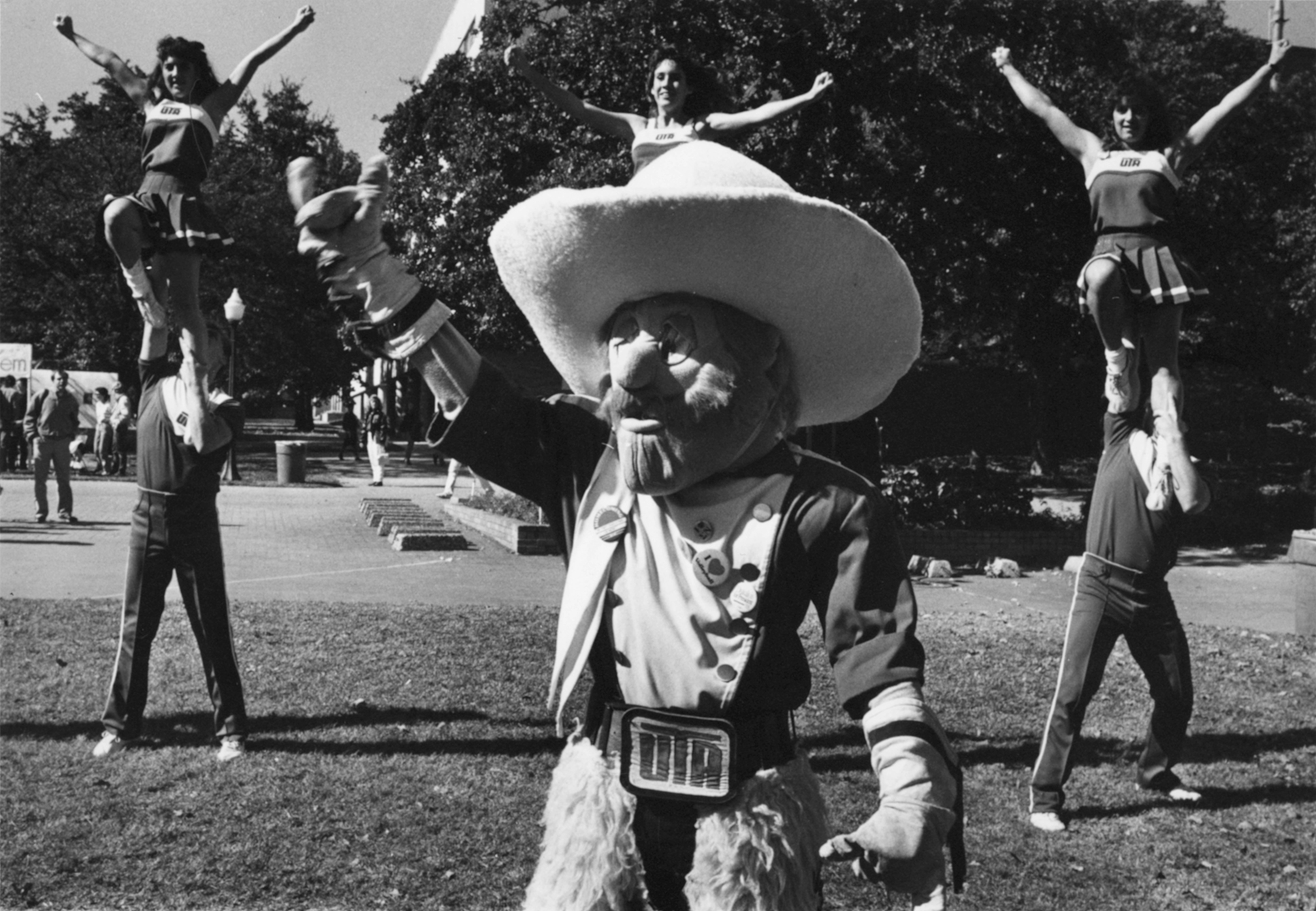 Black and white photo of a mascot wearing a large cowboy hat and western attire with a group of cheerleaders seen in the background.