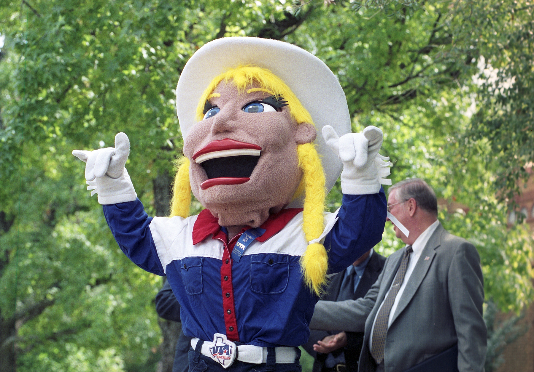 Color photo of a mascot with braided pigtails and a large cowboy hat, which is giving the "Maverick" sign with both of its hands.