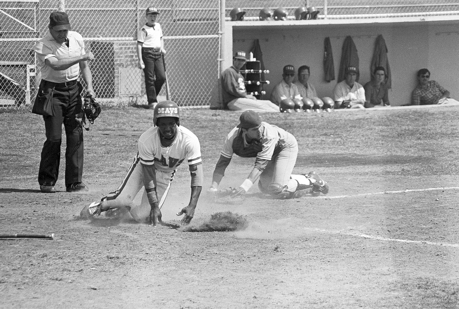 Black and white photo of a man sliding into first base at a baseball game. There is a catcher on his knees attempting to tag the player out. There is an umpire standing just behind them.