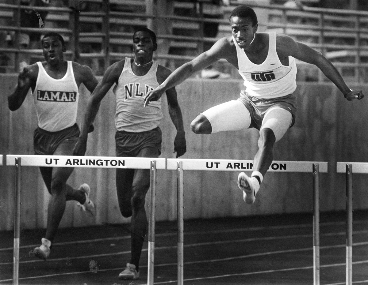 Black and white photo of a track meet with one track member jumping over a hurdle.